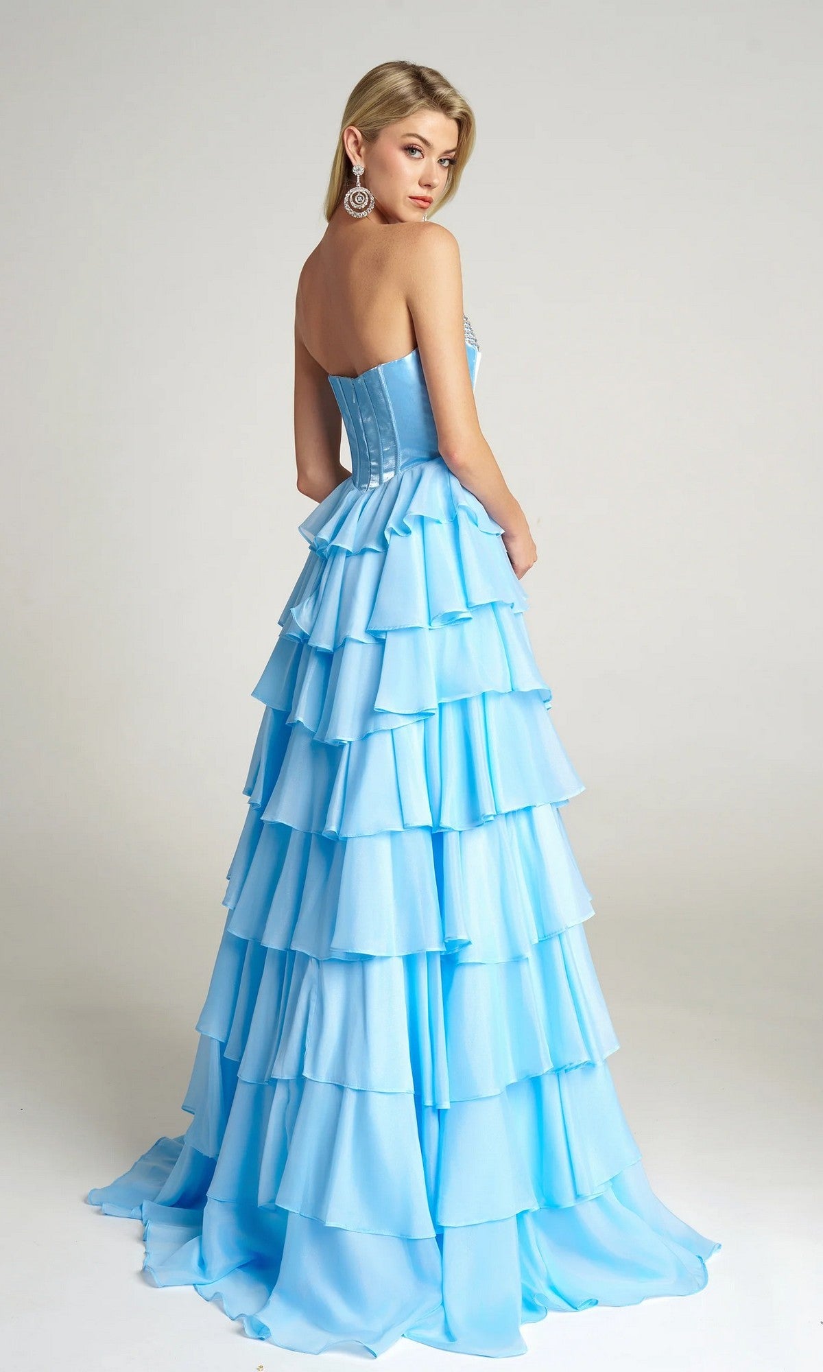A woman wears the Portia and Scarlett PS26024 Long Prom Dress—a strapless, light blue gown with a tiered ruffle skirt and velvet corset bodice—standing slightly turned away from the camera against a plain background.